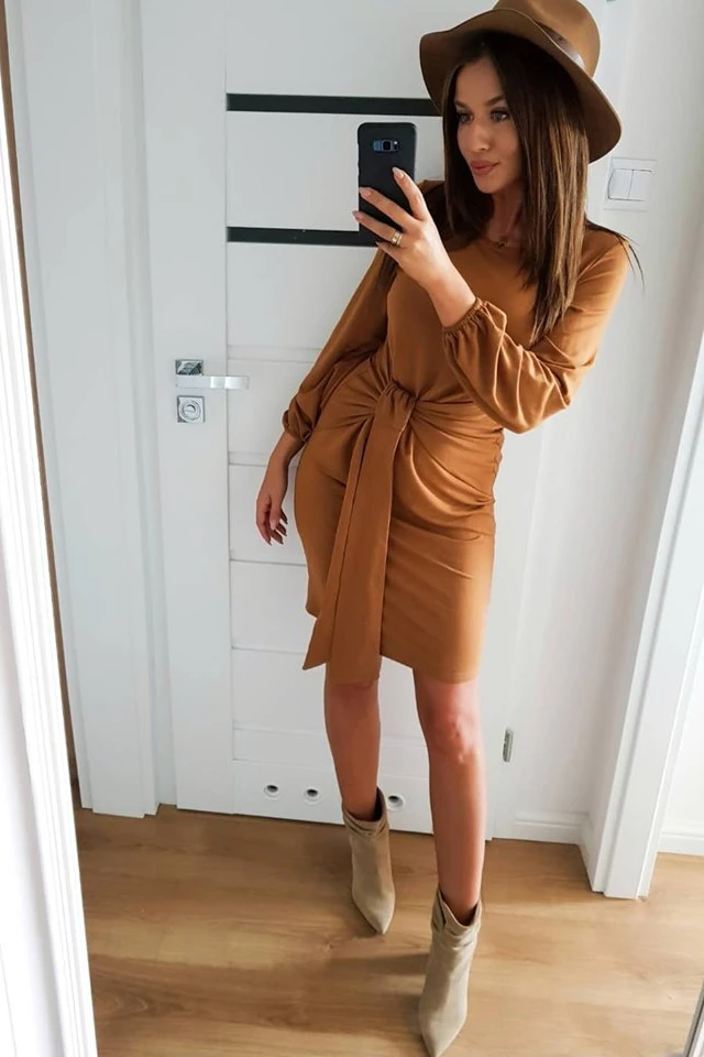 Elegancka sukienka w kolorze karmelowym z długim rękawem i wiązaniem w talii, idealna na jesienne stylizacje. Uzupełniona modnym kapeluszem i beżowymi botkami na obcasie.