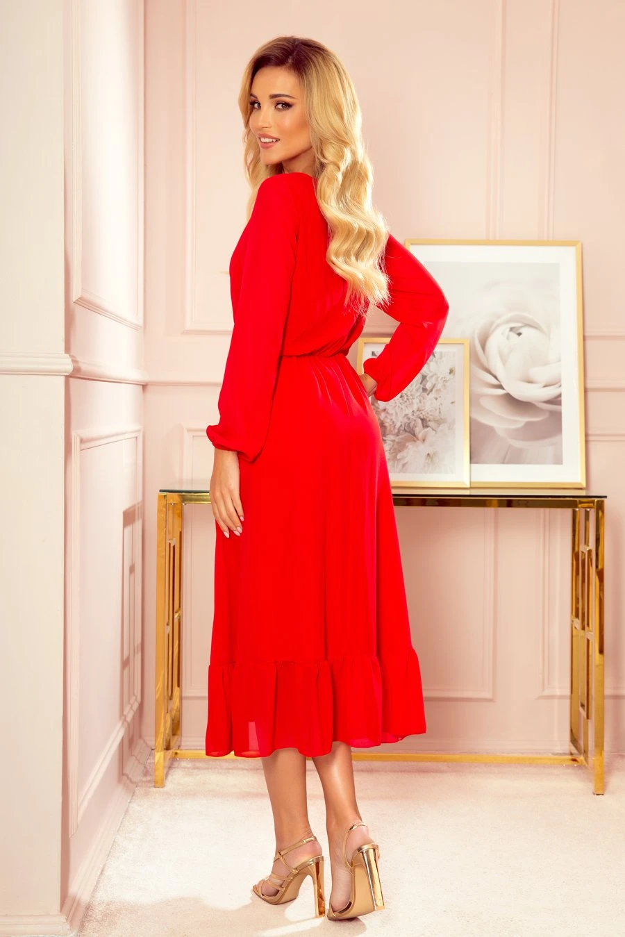 Modelka prezentuje elegancką sukienkę midi w intensywnym czerwonym kolorze, która przyciąga uwagę swoją klasyczną linią i subtelnym falbanowym wykończeniem. Kobieta na zdjęciu emanuje pewnością siebie, podkreślając wyjątkowy styl sukienki midi, idealnej na wyjątkowe okazje.